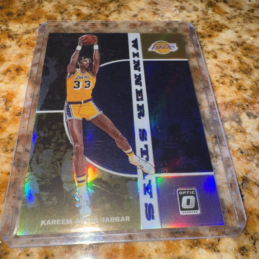 A 2019-20 Panini Donruss Optic Kareem Abdul-Jabbar Lakers card displayed in a clear plastic case on a granite countertop, showing the holographic prizm surface with rainbow light reflections.