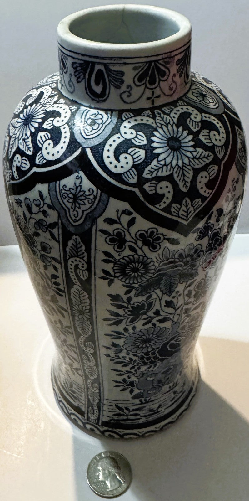 Delft-style blue and white baluster vase standing upright with a quarter at its foot for scale, showing the lambrequin shoulder border and the flowering panels.