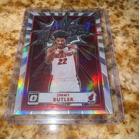 A 2020-21 Panini Donruss Optic Jimmy Butler card in a clear plastic case, displayed on a speckled granite countertop, showing the holographic insert with red borders and the player pointing at the camera.