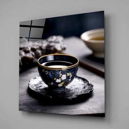 Black decorative tea bowl with gold rim and floral design sits on dark coaster, filled with light liquid, displayed on gray surface.