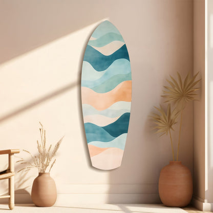 Surfboard-shaped wall art with layered ocean-blue, teal, cream, and peach wavy stripes displayed in minimalist beige room with dried grasses.