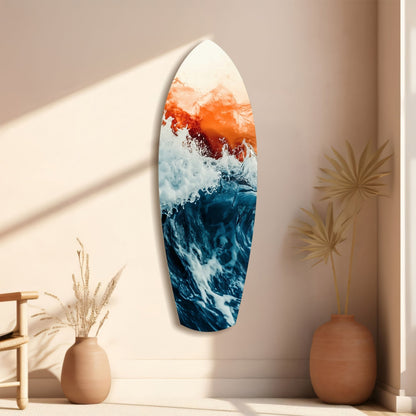 Surfboard-shaped glass wall art with ocean waves in blue, white foam, and orange sunset sky displayed on beige wall with dried plants in terracotta vases.