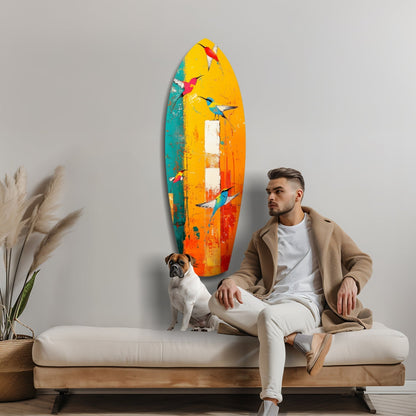 Man seated on white bench beside colorful glass surfboard wall art featuring birds in yellow, orange, teal and red tones, with dog and pampas grass visible.