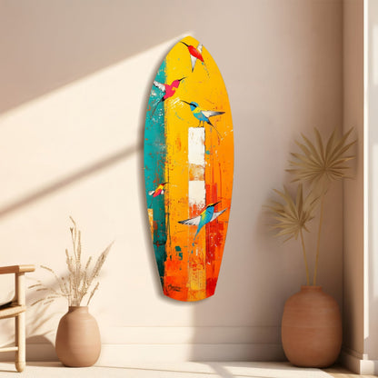 Colorful glass surfboard wall art with painted hummingbirds in yellow, orange, teal, and red tones, displayed in a minimalist room with dried plant decorations.