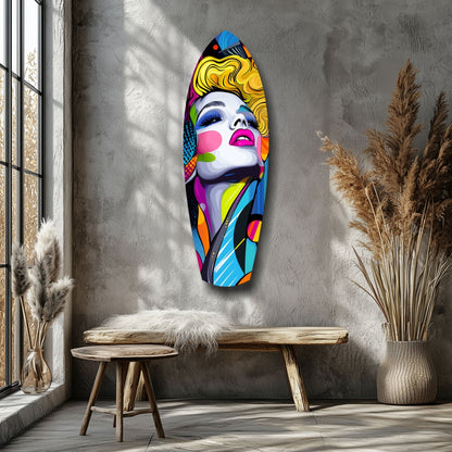 Colorful pop art glass wall sculpture of a woman's face with yellow hair and vibrant geometric patterns, mounted on a gray textured wall above a wooden bench.