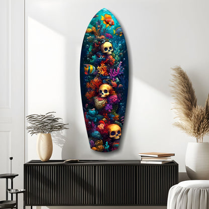 Surfboard-shaped glass wall art displaying golden skulls among vibrant coral, tropical fish, and sea life on dark blue background, mounted in modern white room.