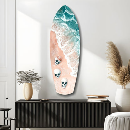 Surfboard-shaped glass wall art featuring aerial ocean waves in teal and white with three skull designs on sandy beach, displayed above black cabinet.