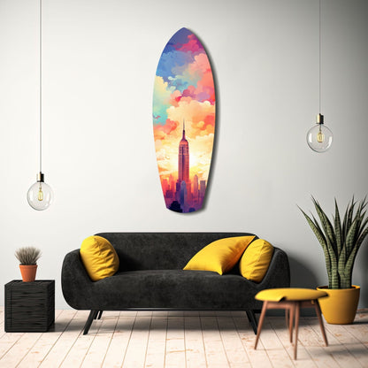 Colorful surfboard-shaped glass wall art depicting Empire State Building at sunset, displayed above black sofa with yellow pillows in modern living room.