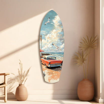Surfboard-shaped glass wall art depicting a vintage red car on a beach with seagulls and lighthouse, displayed on a cream wall between decorative vases.