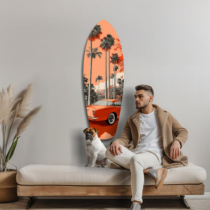 Man and dog sitting on cream sofa beside oval glass wall art featuring red vintage car and palm trees against orange sunset sky.