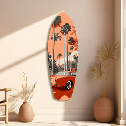 Surfboard-shaped glass wall art featuring a red vintage car and palm trees against an orange sky, displayed on a beige wall with decorative vases.
