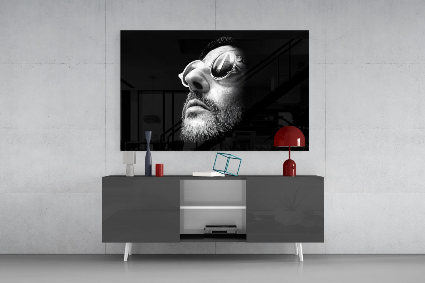 Black and white portrait of bearded man wearing oval sunglasses displayed on wall-mounted television above gray console table with red lamp and accessories.