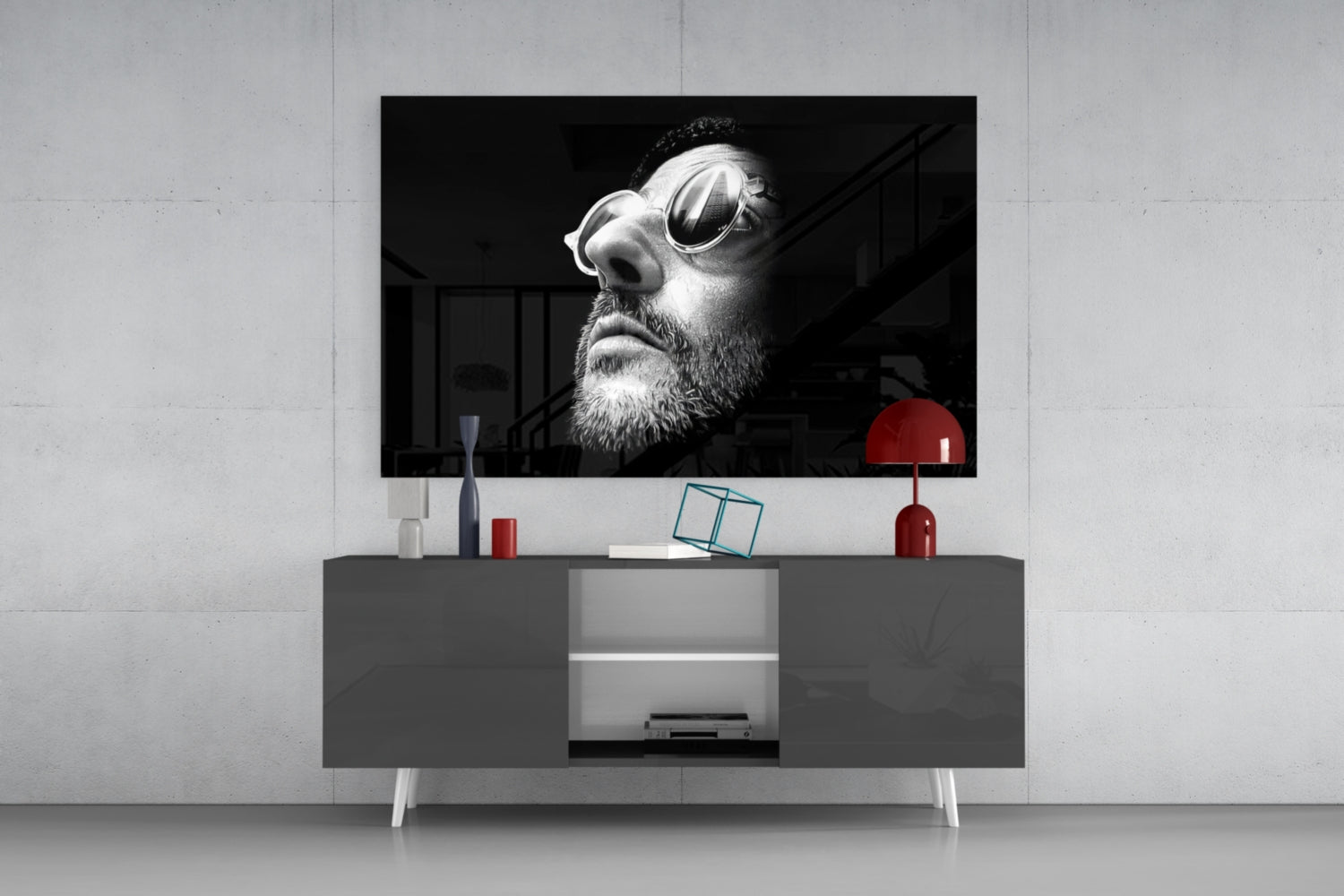 Black and white portrait of bearded man wearing oval sunglasses displayed on wall-mounted television above gray console table with red lamp and accessories.