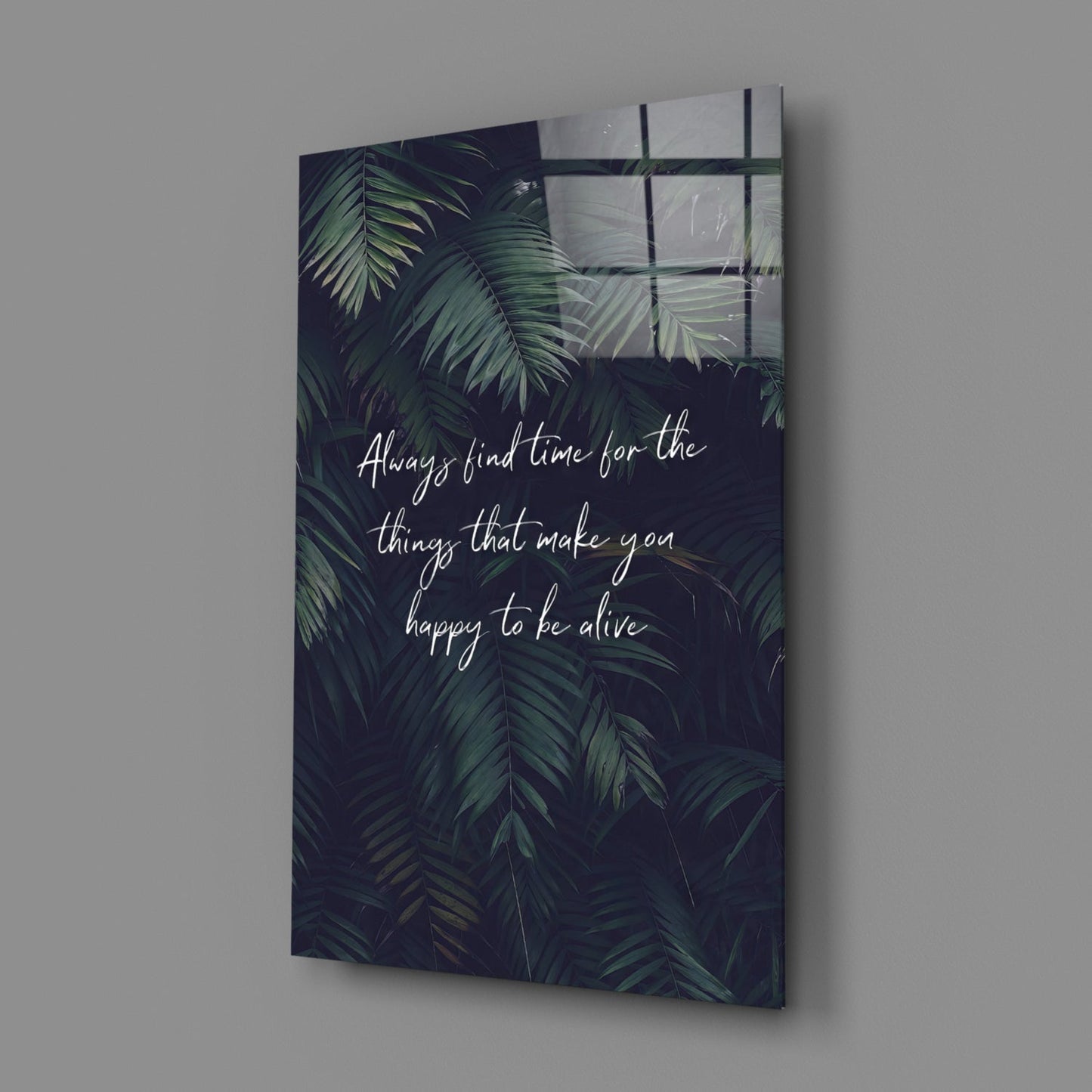 Glass wall art with dark tropical foliage background and white handwritten text reading "Always find time for the things that make you happy to be alive," displayed at an angle against a gray wall.