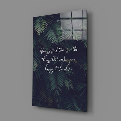 Glass wall art with dark tropical foliage background and white handwritten text reading "Always find time for the things that make you happy to be alive," displayed at an angle against a gray wall.