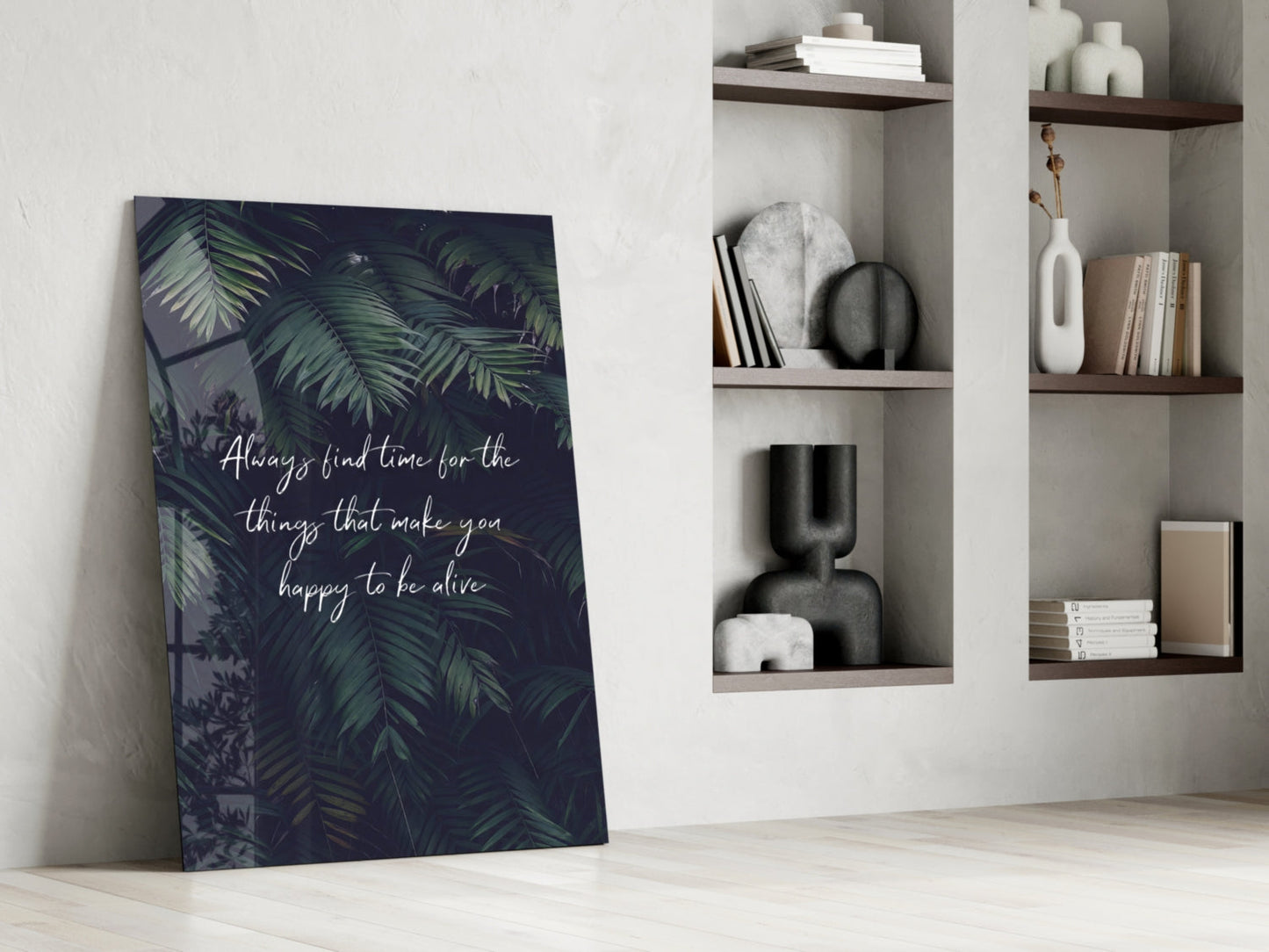 Glass wall art with dark tropical foliage and white script text leaning against white wall beside wooden shelves with decorative objects.