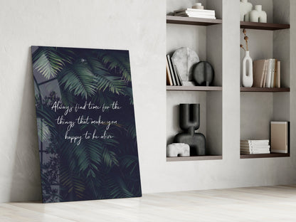 Glass wall art with dark tropical foliage and white script text leaning against white wall beside wooden shelves with decorative objects.