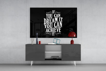 Black glass wall art with white inspirational text mounted above gray media console in modern living room setting with red lamp and decorative accessories.