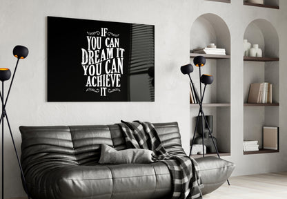 Black glass wall art with white motivational text hung above a gray leather sofa in a modern living room setting.