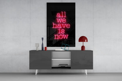 Pink neon glass wall art reading "all we have is now" displayed above a dark gray media console with decorative accessories.