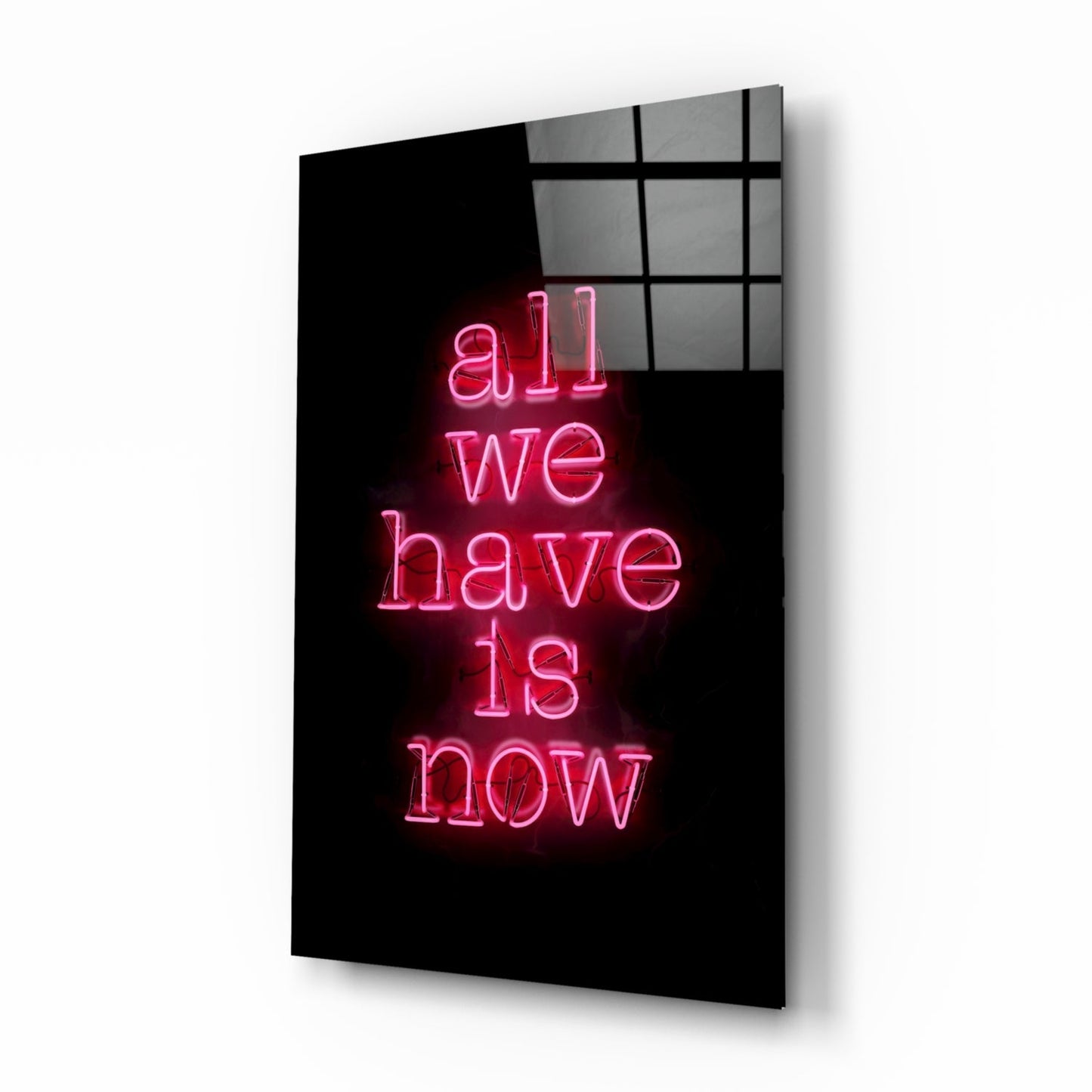 Pink neon text reading "all we have is now" glows against black background on angled glass panel with gridded window panes visible above.