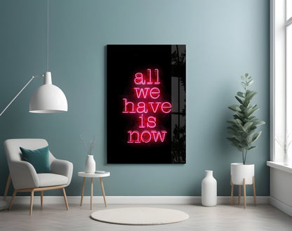 Neon glass wall art displaying "all we have is now" in pink lettering mounted in a minimalist room with teal walls, white furniture, and potted plants.