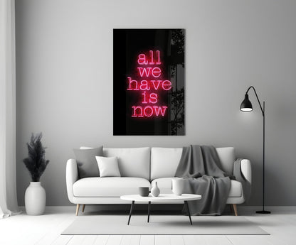 Pink neon text reading "all we have is now" on black glass mounted above white sofa in modern living room interior.