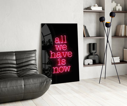 Pink neon "all we have is now" text on black glass leaning against white wall in modern living room setting.