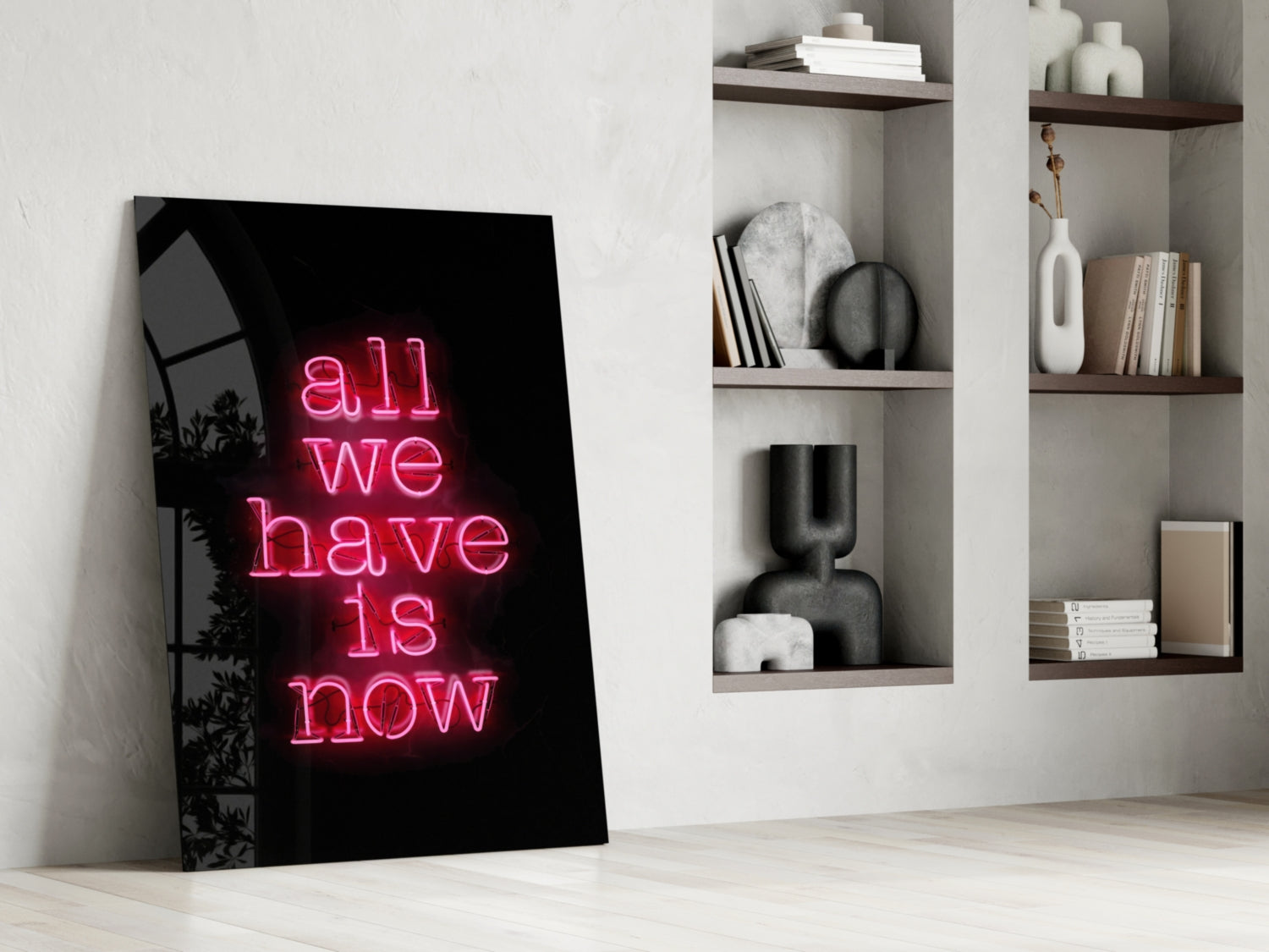 Black glass wall art with pink neon-style "all we have is now" text leaning against white wall beside shelving unit.