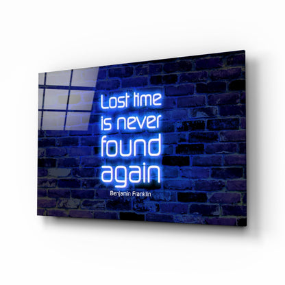 Blue neon sign on dark brick wall displaying "Lost time is never found again" quote by Benjamin Franklin, mounted on glass panel with window reflection.