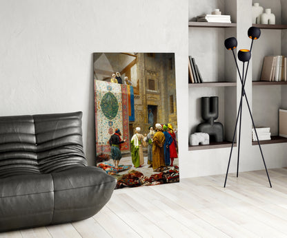 Glass wall art depicting a Middle Eastern bazaar scene with figures in colorful robes examining ornate carpets and textiles, displayed in a modern living room setting.