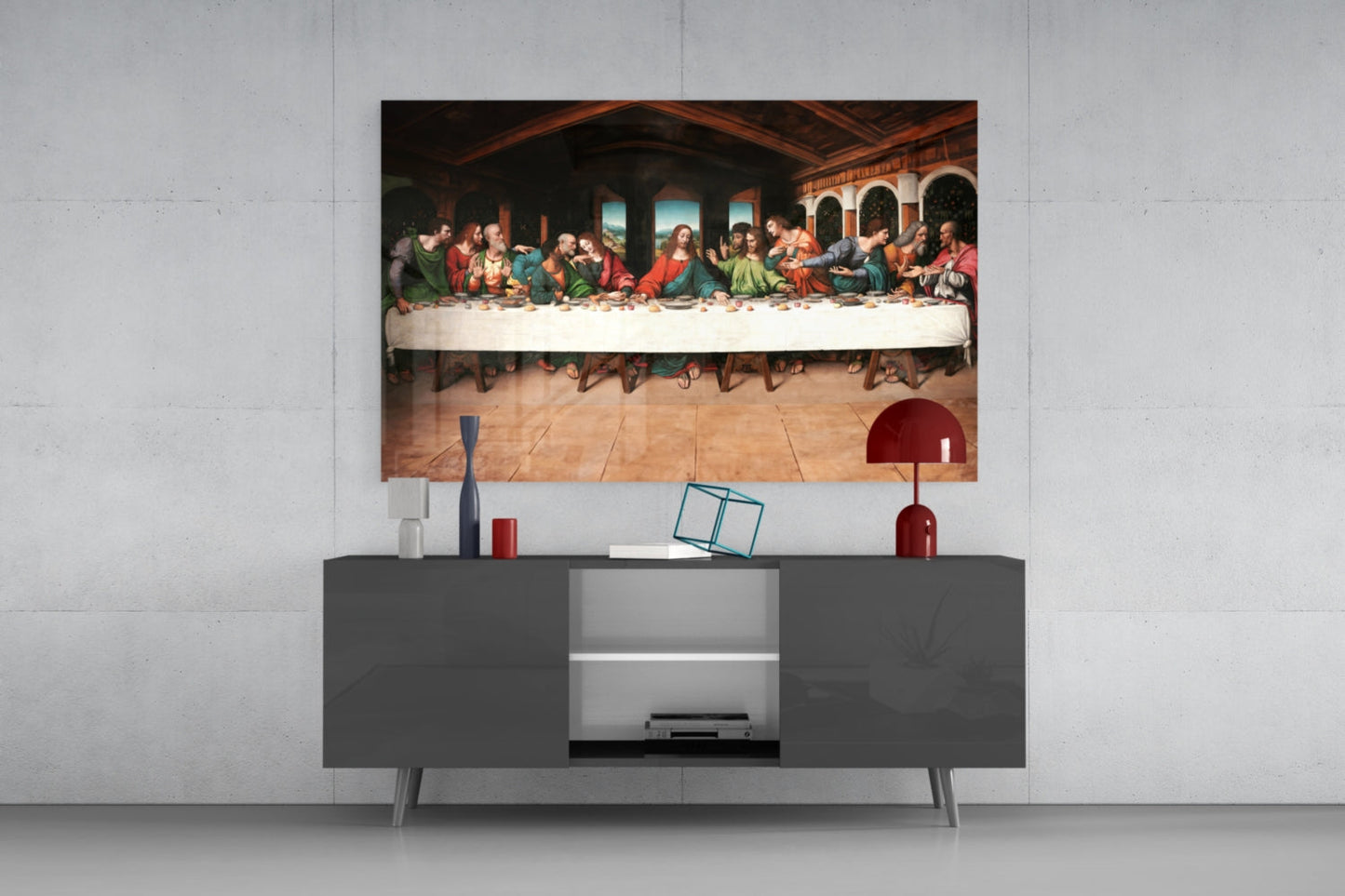 Glass wall art depicting the Last Supper displayed above a dark gray media console with red decorative accessories.
