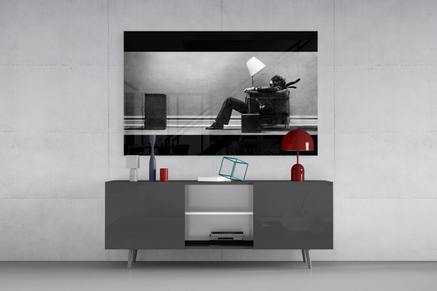 Glass wall art mounted above gray media console displaying black-and-white photograph of man in chair with white lamp, red decorative objects on shelf below.