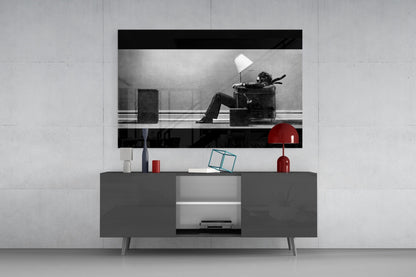 Glass wall art mounted above gray media console displaying black-and-white photograph of man in chair with white lamp, red decorative objects on shelf below.