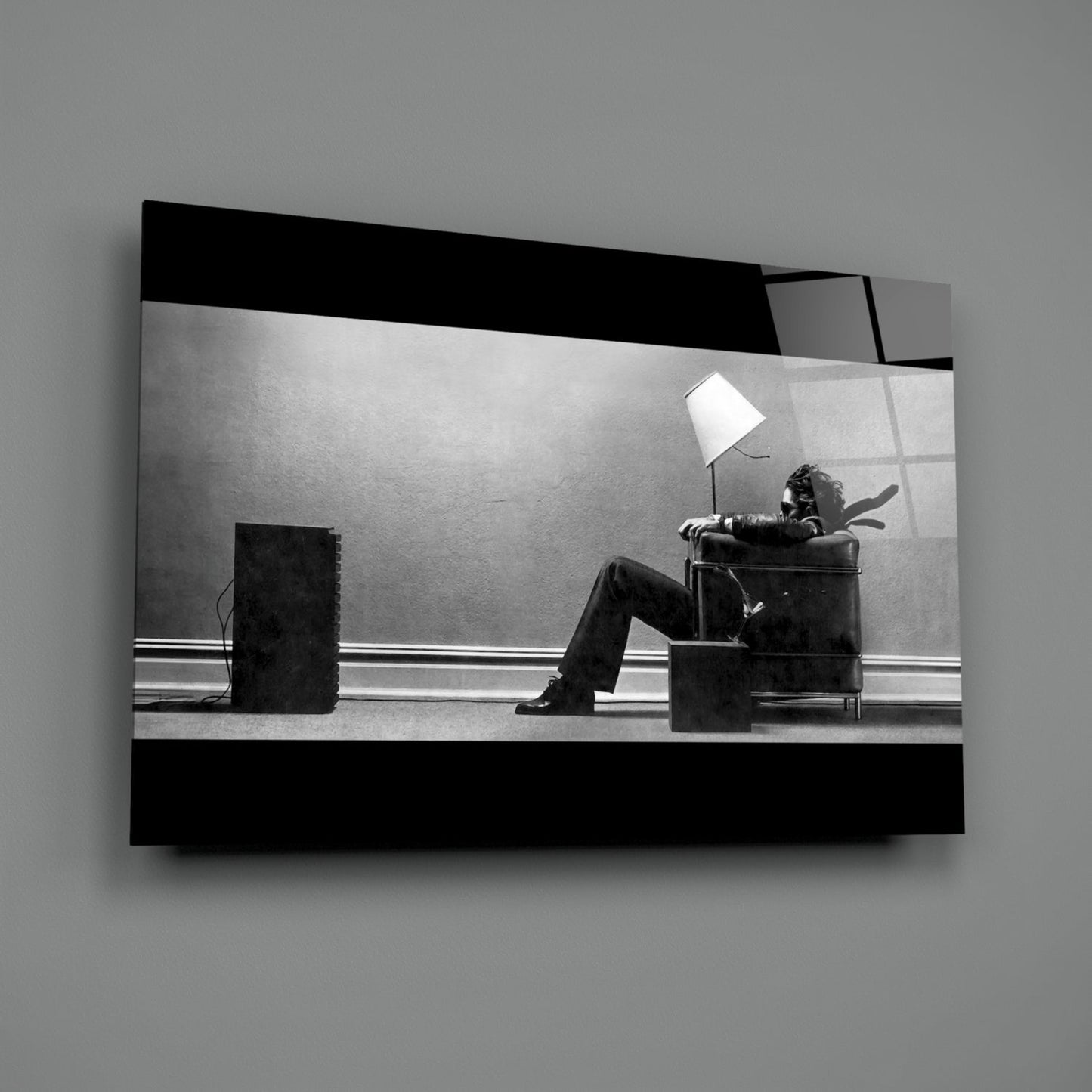 Black and white glass wall art showing a figure seated in a chair beneath a lamp, displayed at an angle against gray background.