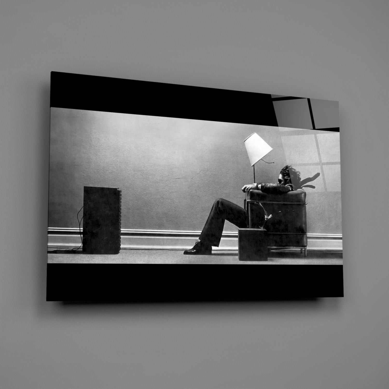 Black and white glass wall art showing a figure seated in a chair beneath a lamp, displayed at an angle against gray background.