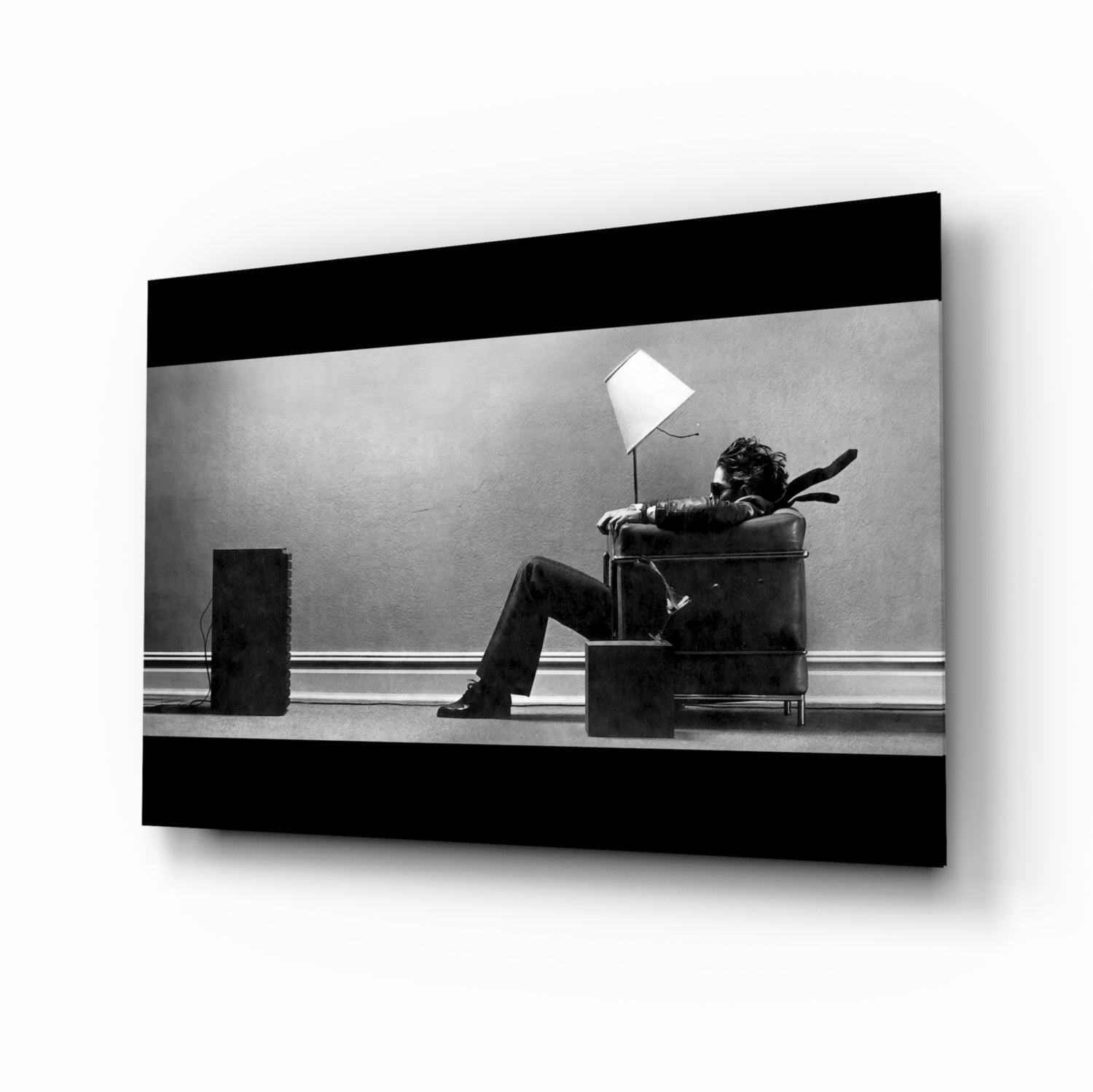 Black and white glass wall art showing a person sitting in a leather chair with a lamp, displayed at an angle with shadow.