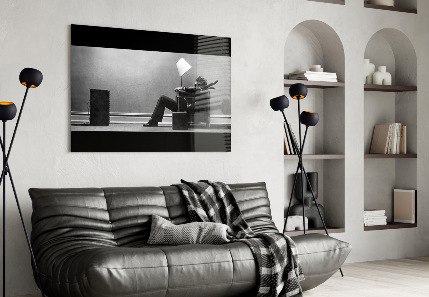 Black and white glass wall art depicting a silhouetted figure sitting in a chair with a lamp, displayed above a black leather sofa in a modern living room setting.