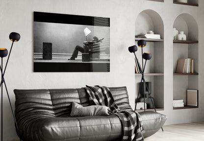 Black and white glass wall art depicting a silhouetted figure sitting in a chair with a lamp, displayed above a black leather sofa in a modern living room setting.