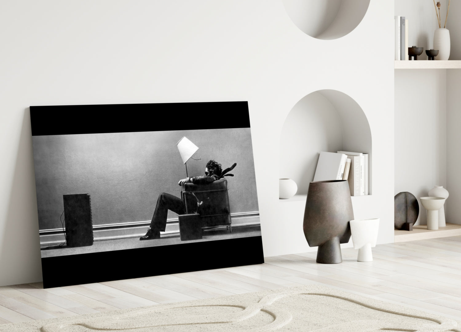Black and white framed glass art depicting a person sitting in a chair under a lamp, displayed leaning against a white wall in a minimalist interior setting.