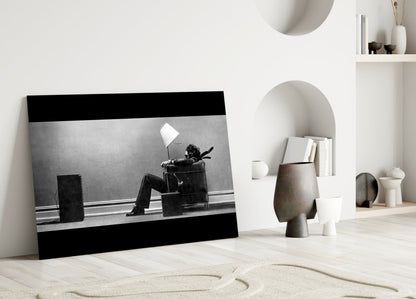 Black and white framed glass art depicting a person sitting in a chair under a lamp, displayed leaning against a white wall in a minimalist interior setting.