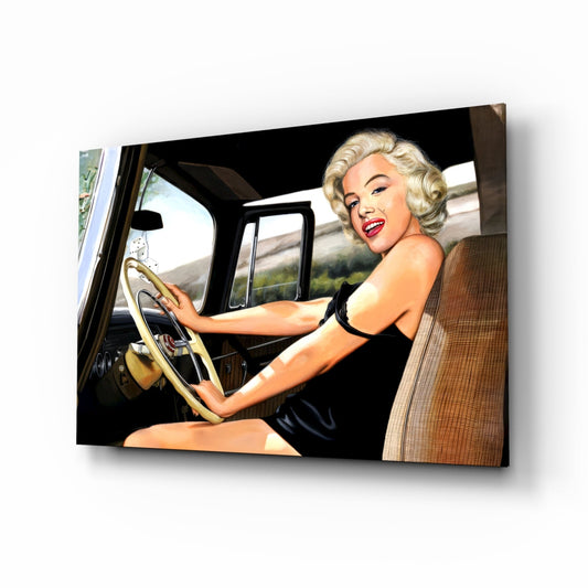 Blonde woman with red lipstick at steering wheel of vintage car, wearing black dress, smiling at camera.