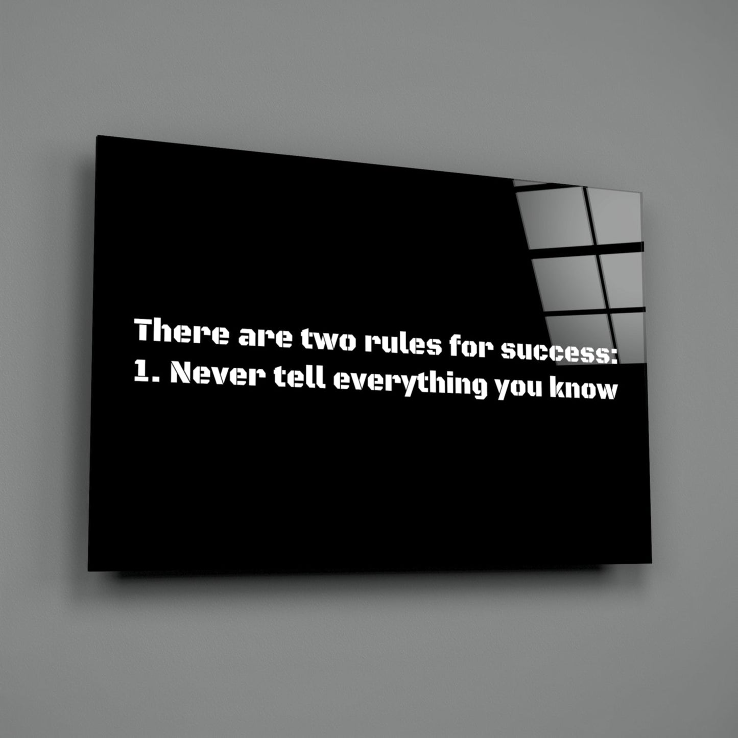 Black glass wall art panel with white text reading "There are two rules for success: 1. Never tell everything you know" displayed at an angle against gray wall.