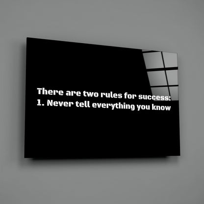 Black glass wall art panel with white text reading "There are two rules for success: 1. Never tell everything you know" displayed at an angle against gray wall.