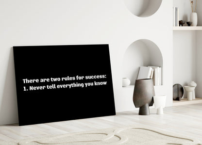 Black glass wall art displaying white text "There are two rules for success: 1. Never tell everything you know" leaning against white interior wall with shelving and decorative objects.