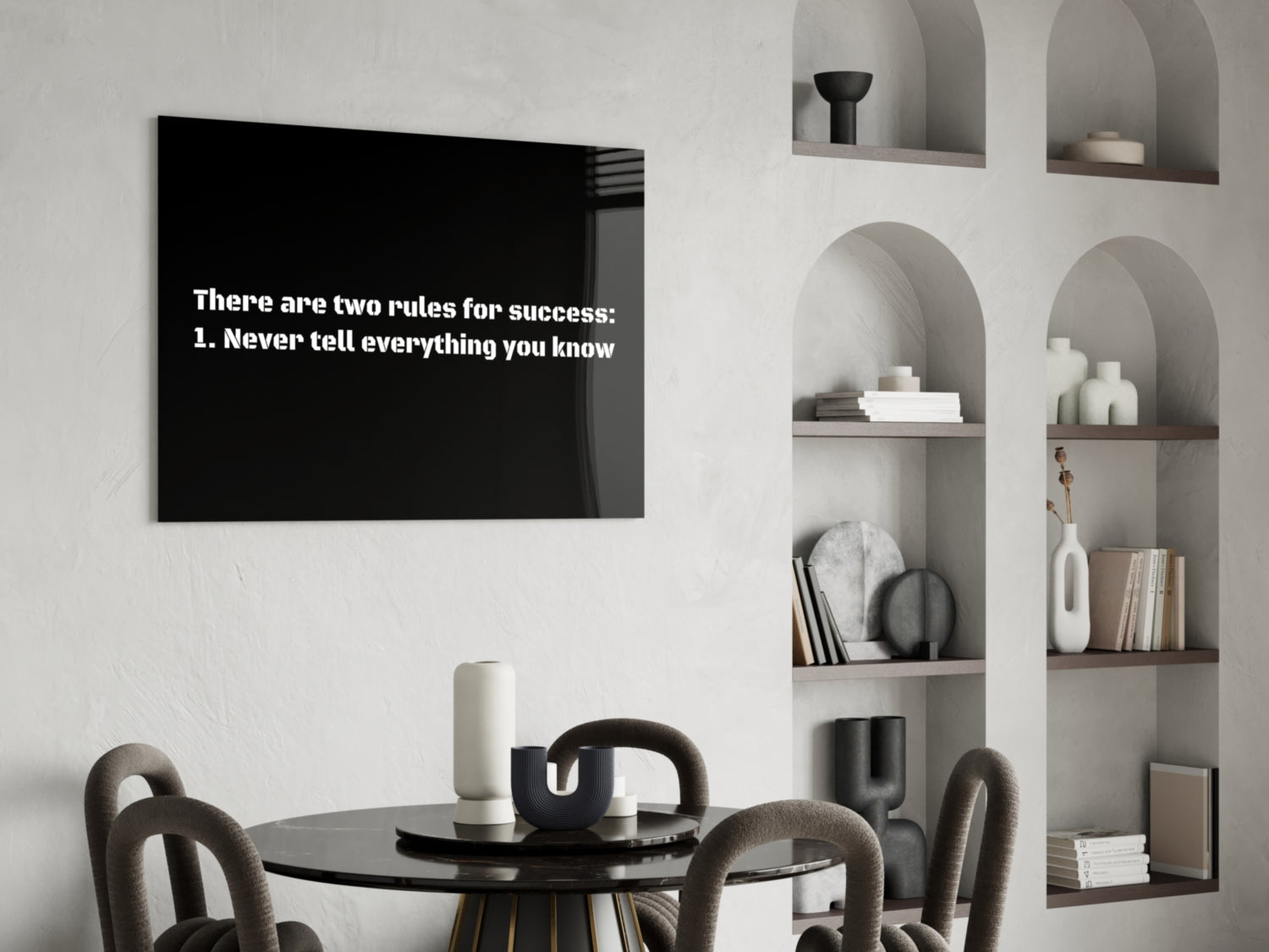 Black glass wall art displaying white text "There are two rules for success: 1. Never tell everything you know" hung above a round dining table in a minimalist room.
