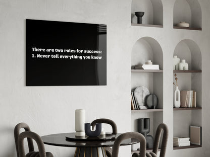 Black glass wall art displaying white text "There are two rules for success: 1. Never tell everything you know" hung above a round dining table in a minimalist room.