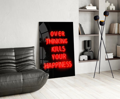 Red neon "OVER THINKING KILLS YOUR HAPPINESS" text on black glass leaning against white wall in modern living room setting.