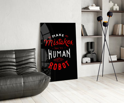 Black glass wall art with red and white text reading "Make Mistakes, You're Human Not a Robot" displayed in modern living room next to gray sofa.