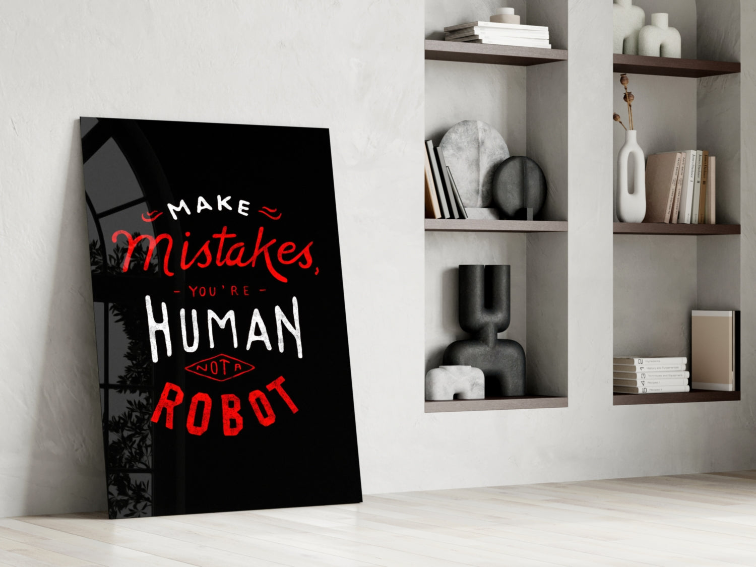 Black glass wall art with red and white text reading "MAKE MISTAKES, YOU'RE HUMAN NOT A ROBOT" leaning against white wall beside shelving.